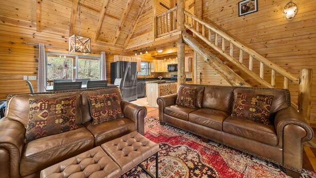 Ein Holzblockhaus Innenansicht mit einem Wohnzimmer, das Ledersofas und einen gemusterten Teppich sowie eine Küchenzeile und eine Treppe zeigt.