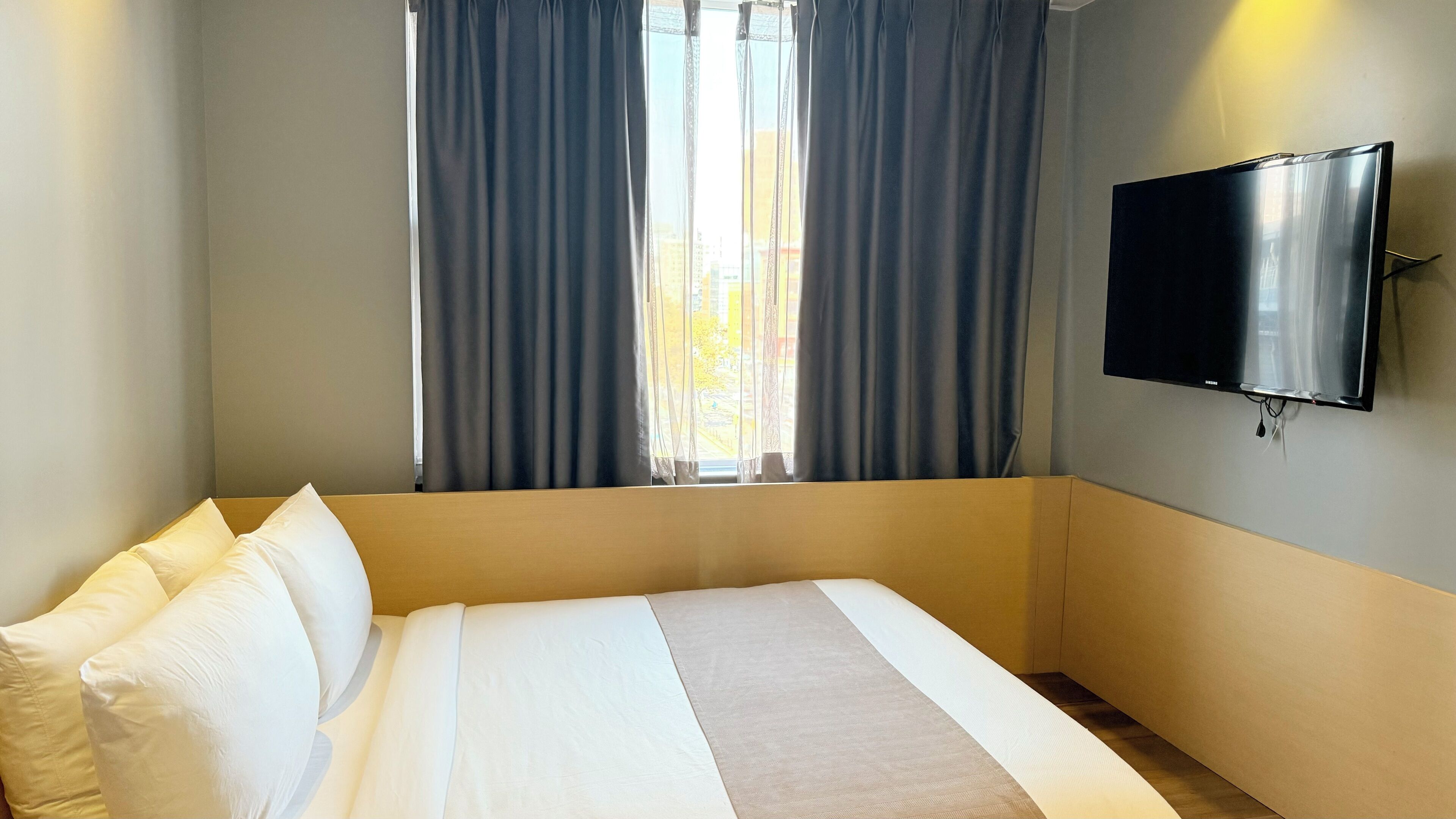 A hotel room with a bed, a flat-screen TV mounted on the wall, and a window with curtains.