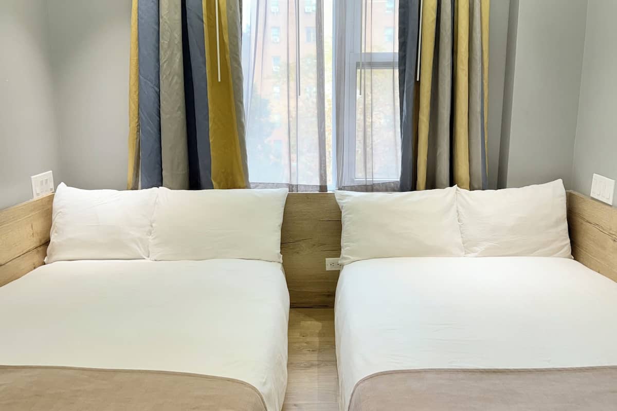 Una cama doble con ropa de cama blanca en una habitación con cabecera de madera, una ventana con cortinas y un interruptor de luz en la pared.