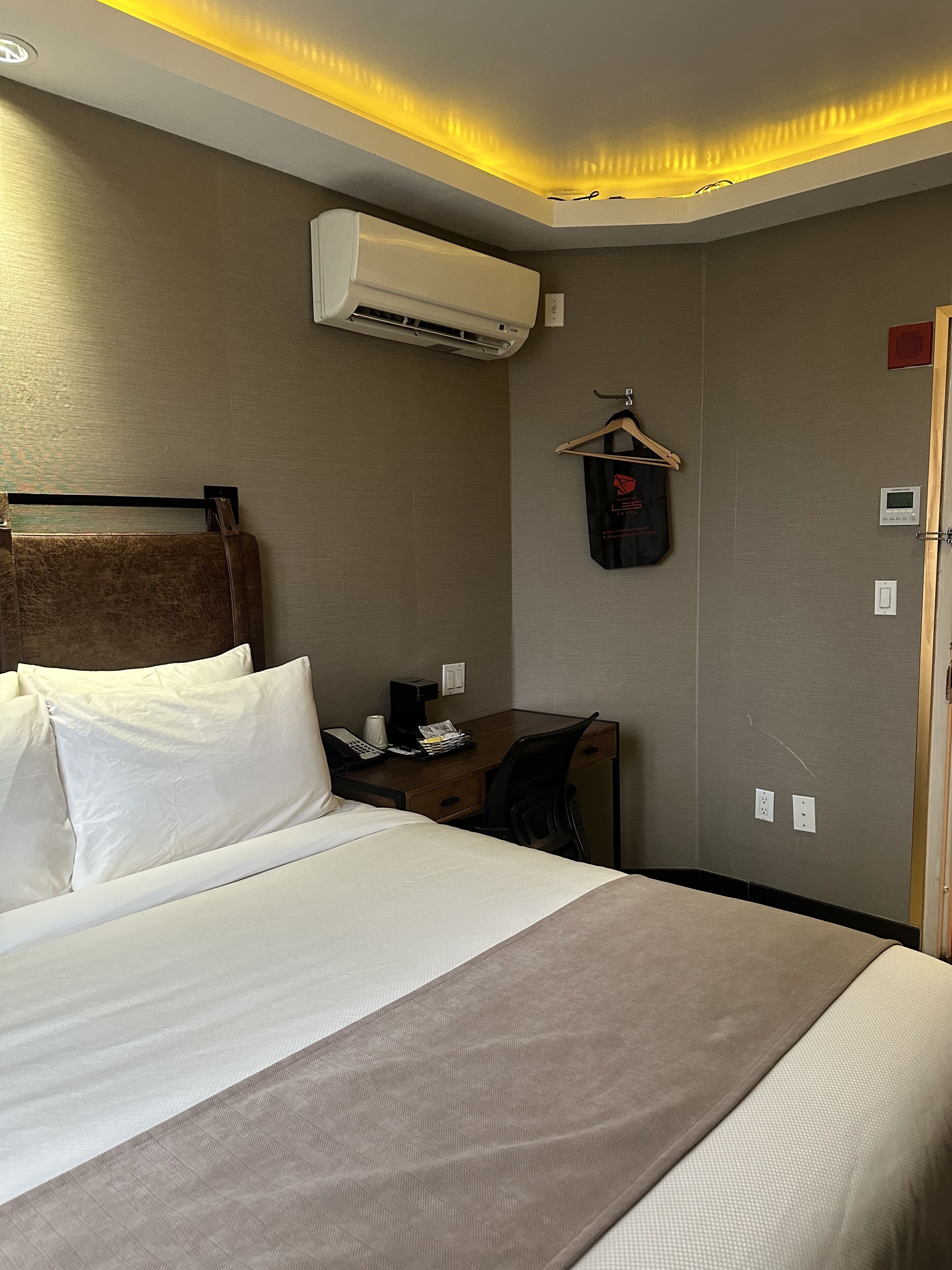 Ein Hotelzimmer mit Bett, Schreibtisch, Stuhl, Klimaanlage, Kleiderhaken und einer aufhängbaren Tasche.