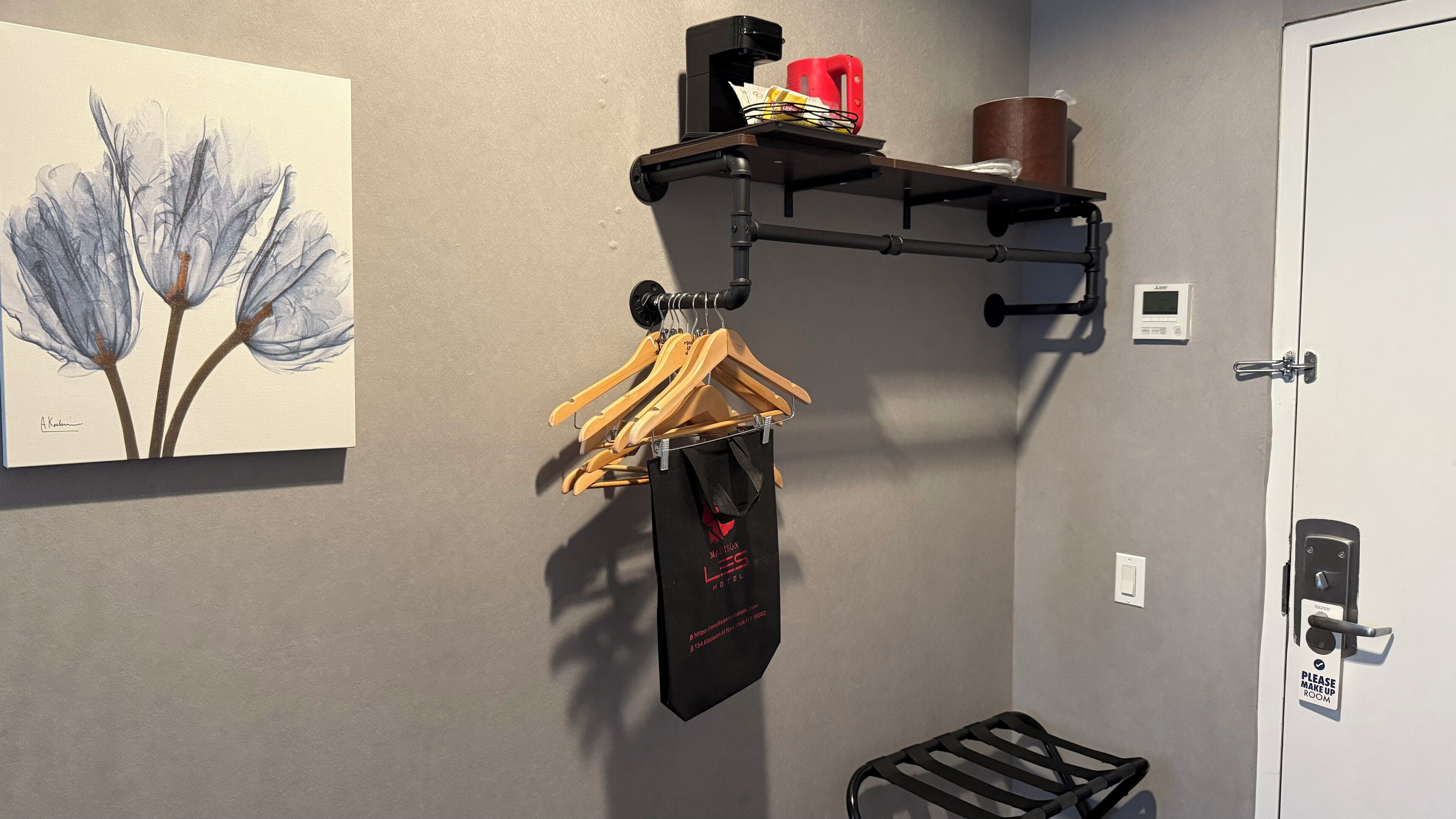 A hotel room with a black shelf holding a red cup, a brown container, and a small white device. A black tie hangs from a wooden hanger, and a black bag is attached to the same hanger. A painting of blue flowers is on the wall.
