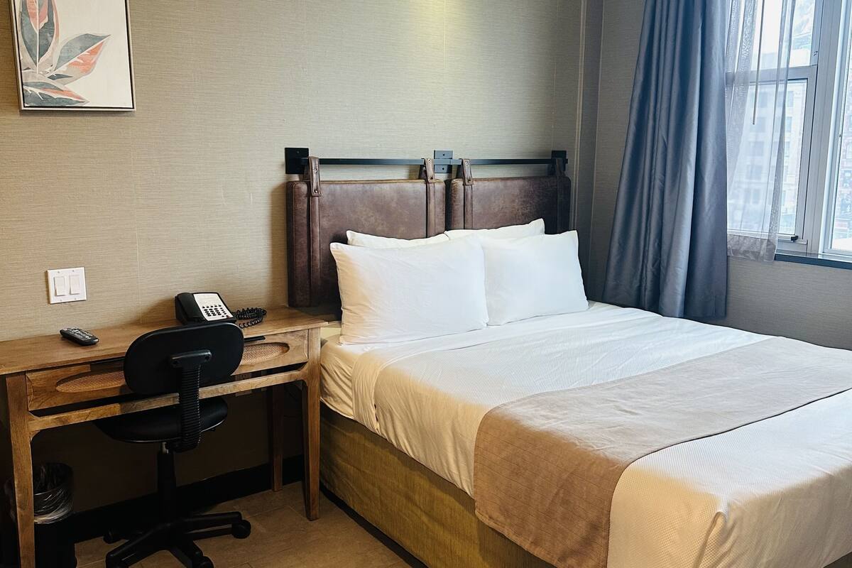 Habitación de hotel con una cama grande, un escritorio con silla, un teléfono y una ventana con cortinas.