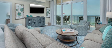 Un salon spacieux avec un grand canapé, une table basse et un téléviseur à écran plat. Il y a un balcon avec vue sur l’océan.