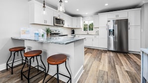 Una cocina moderna con armarios blancos, un refrigerador de acero inoxidable, una isla de granito con taburetes y iluminación colgante.