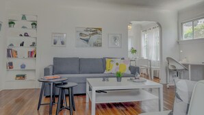 Ein modernes Wohnzimmer mit einem grauen Sofa, einem weißen Couchtisch und schwarzen Hocker. An den Wänden befinden sich Bücherregale mit Dekoration und Gemälde.