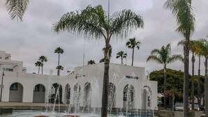 Ein öffentlicher Brunnen mit mehreren Wasserstrahlen, umgeben von Palmen und einem modernen Gebäude mit Rundbogenfenstern.