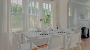 Ein Essbereich mit einem weißen Tisch, gedeckt für vier Personen, umgeben von weißen Stühlen, großen Fenstern mit durchscheinenden Vorhängen und Blick auf Grünfläche.