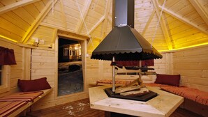 A log cabin interior with a central wood-burning stove, wooden benches, and a cozy ambiance. - Kärkiniemi (Tuusniemi)