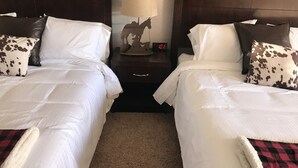 A hotel room with two beds, each with white bedding and patterned pillows. There is a nightstand with a lamp and a clock between the beds. - Country West Motel (Rock Springs)