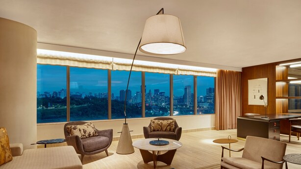 Une chambre d’hôtel moderne dotée d’une grande fenêtre donnant sur la ville, d’un coin salon confortable avec une table ronde et d’un bureau avec une lampe.
