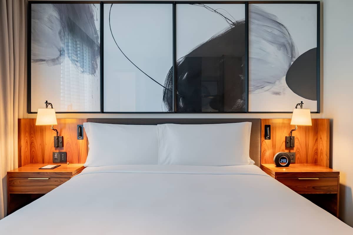 Hotelkamer met een groot abstract schilderij boven het bed, twee nachtkastjes met lampen, een klok en een telefoon op het linker nachtkastje.