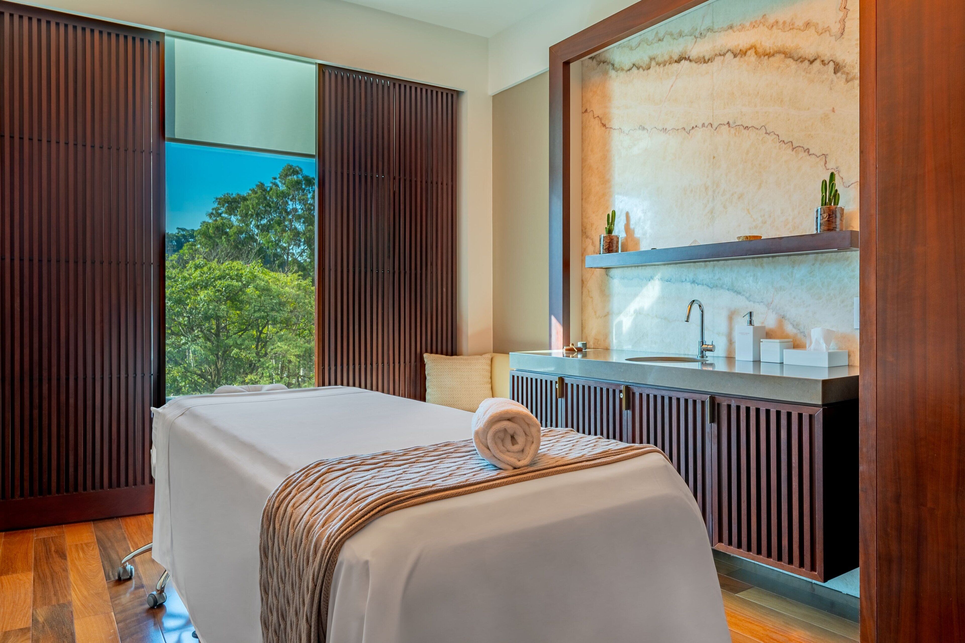Habitación de hotel moderna con una cama grande, suelo de madera, vista a árboles a través de una puerta corrediza y una pared de mármol con lavamanos y espejo.
