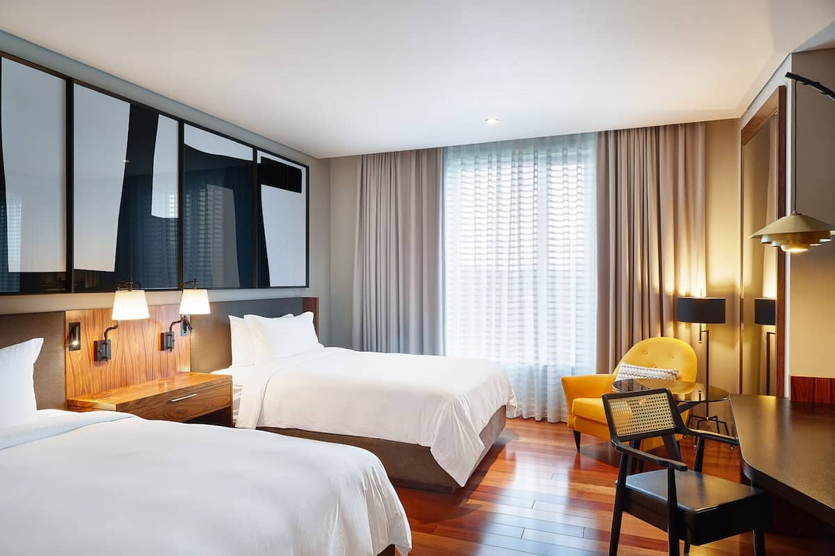 Een hotelkamer met twee bedden, een bureau, een stoel en een groot raam met gordijnen.