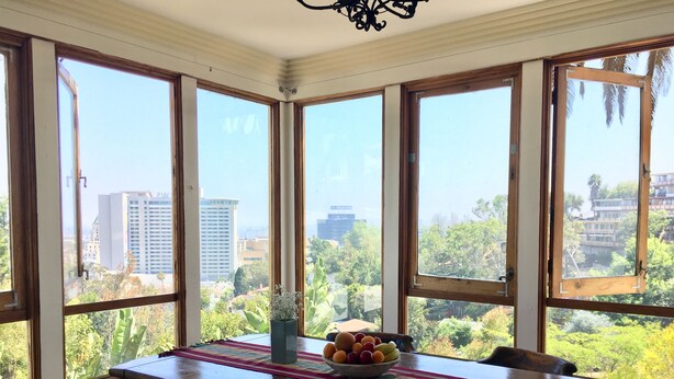 A dining area with a wooden table, chairs, and a fruit bowl. Large windows offer a view of buildings and trees. - Classic Hollywood Hills Living with Amazing Views ~1200sf (Los Angeles)