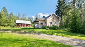 Ein zweistöckiges Haus mit einem großen Vorgarten, einem roten Bauernhaus und bewaldeter Landschaft im Hintergrund.