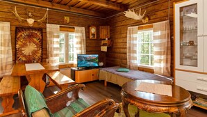 Ein gemütliches Blockhaus-Interieur mit Holzwänden, Holzbalkendecke, einem Bett, einem Fernseher, einem Esstisch und einem Kamin.
