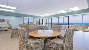 A spacious room with a round table and wicker chairs, large windows with a view of the ocean, and a ceiling with recessed lighting. - Phoenix East 1003 - Beautiful 2 BR Condo (Orange Beach)