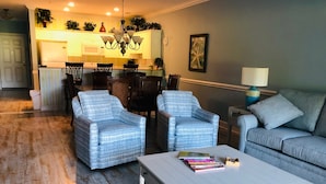 A living room with a white coffee table, two blue armchairs, a grey sofa, and a dining area with a chandelier.