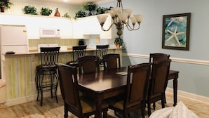 A dining area with a wooden table and chairs, a chandelier, and a kitchen in the background.
