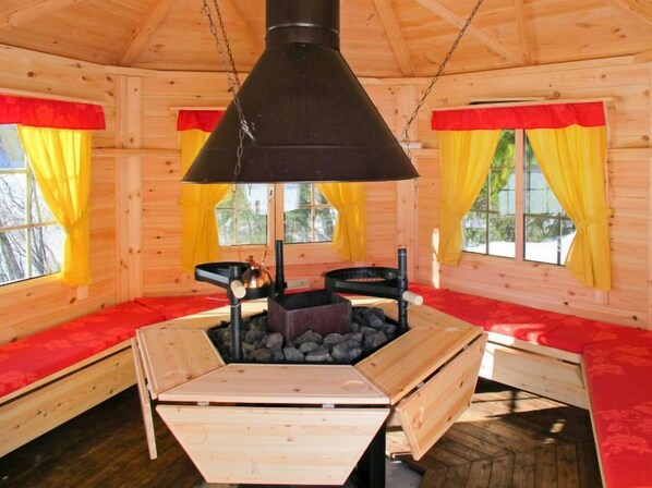 Interior acogedor de una cabaña con chimenea central, paredes de madera y cortinas rojas y amarillas.