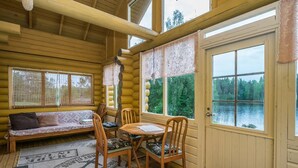 A log cabin interior with a wooden table and chairs, a sofa, and large windows overlooking a lake.