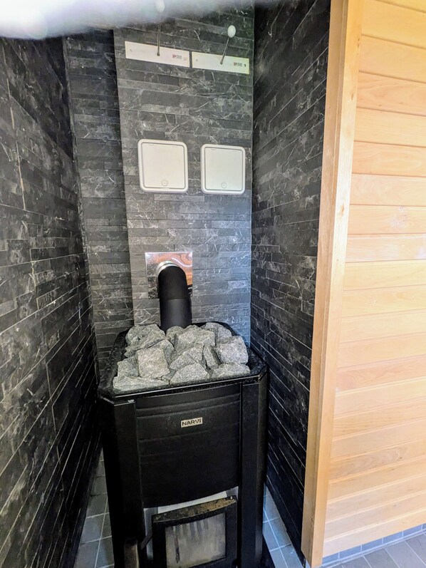 A compact room with a black marble wall, a black heater with a stone facade, and a small black cabinet labeled "NARVI". - Joutsenranta (Tuusniemi)