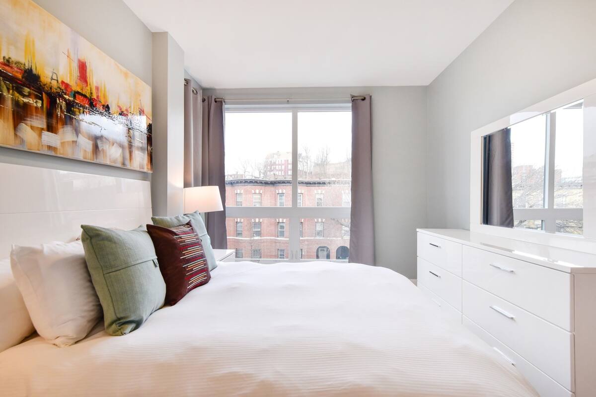 A hotel room with a large bed, a white dresser, a window with curtains, and a cityscape view.