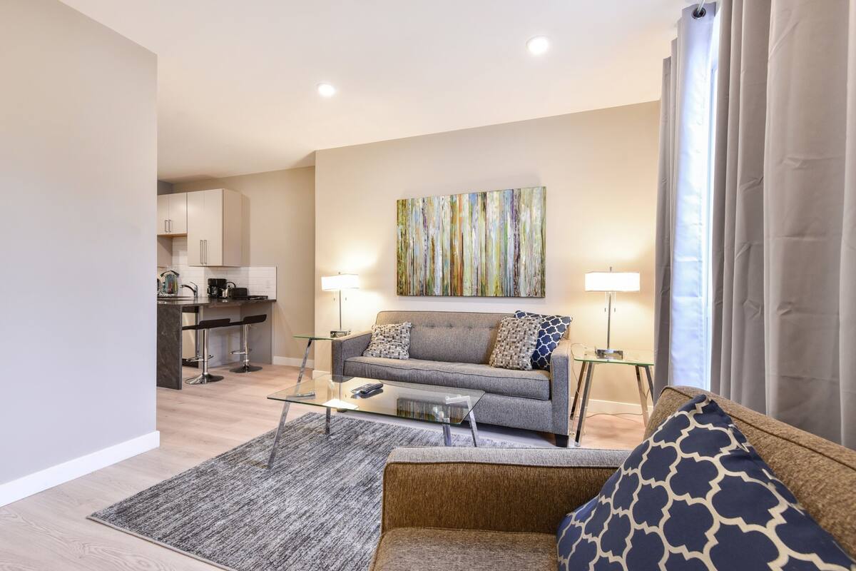 A modern living room with a grey sofa, a coffee table, and a patterned rug. There is a kitchen area in the background with a bar and a dining table.