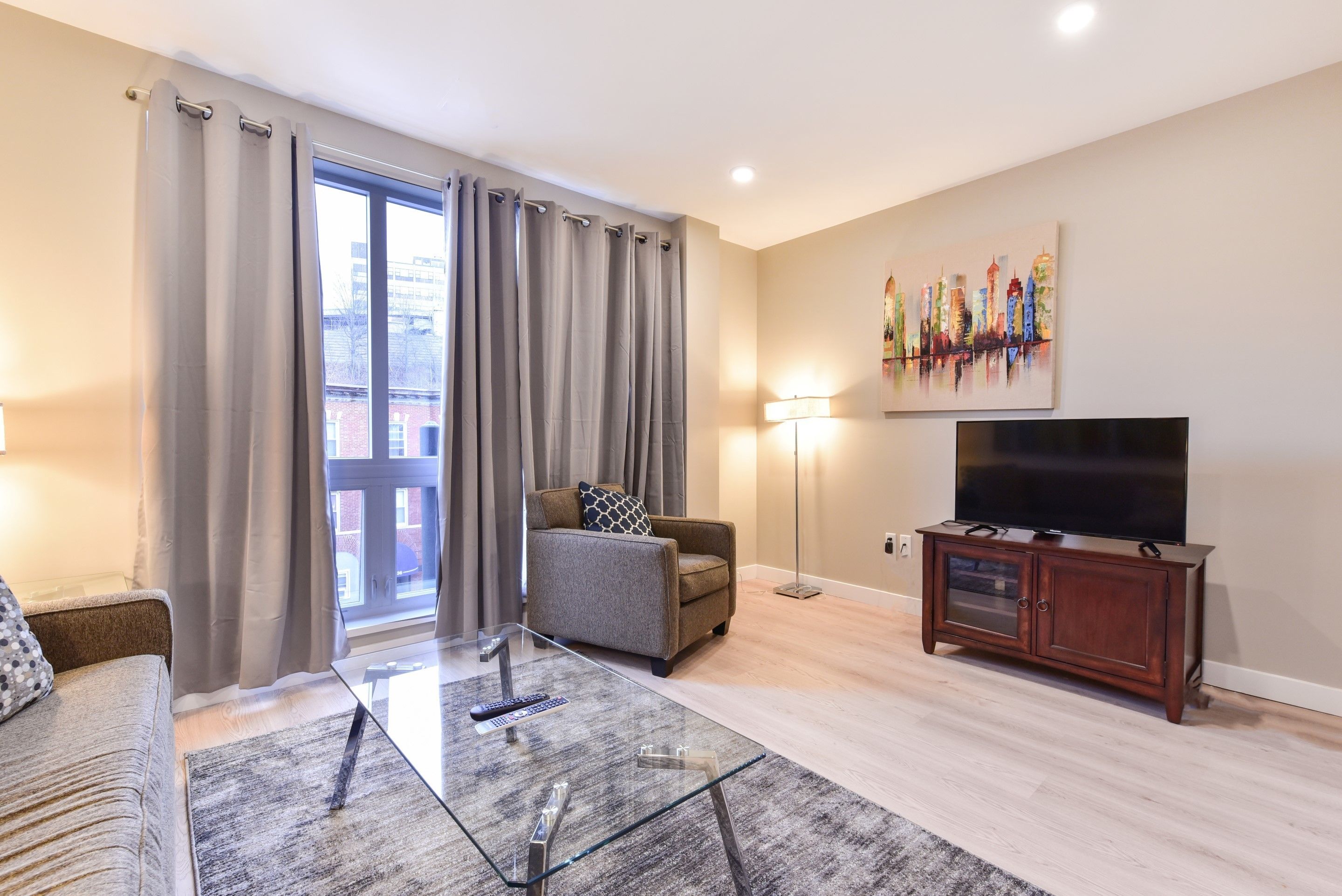 Una sala de estar con sofá, sillón, mesa de centro de vidrio, mueble para televisor y cortinas.