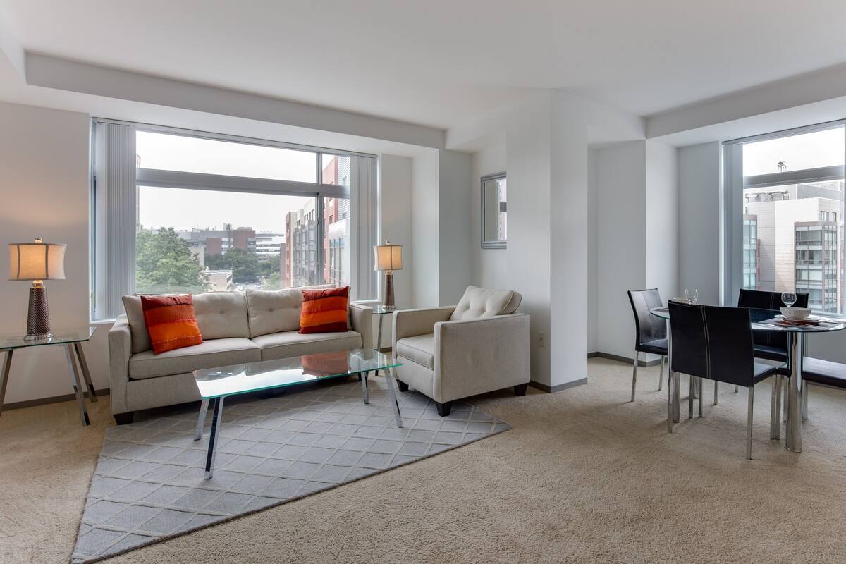 A modern living room with a sofa, armchair, glass coffee table, and dining table with chairs. Large windows provide views of the city.