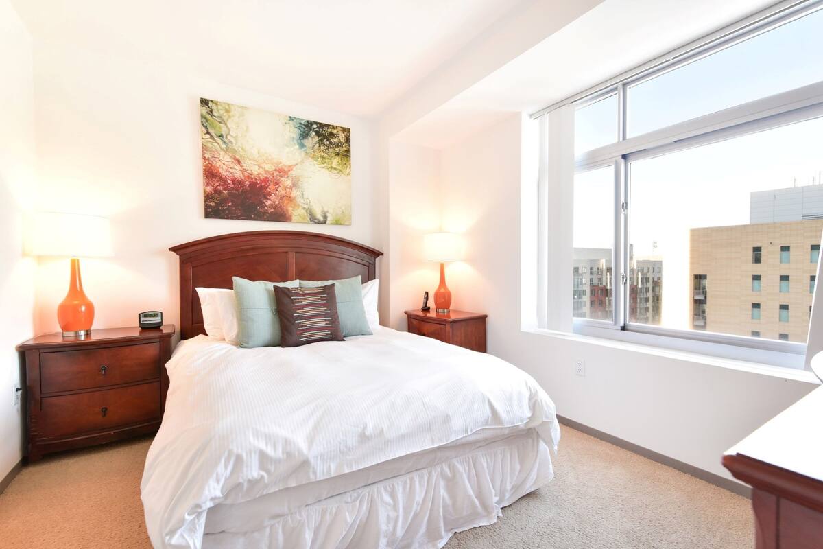 A bedroom with a large bed, two bedside tables with lamps, a painting on the wall, and a window with a city view.