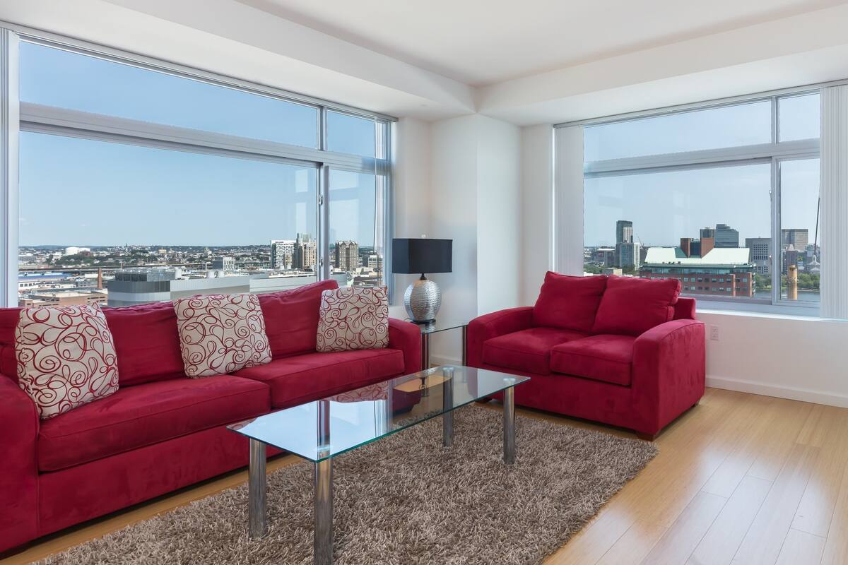 A modern living room with two red sofas, a glass coffee table, and a view of a cityscape through large windows.