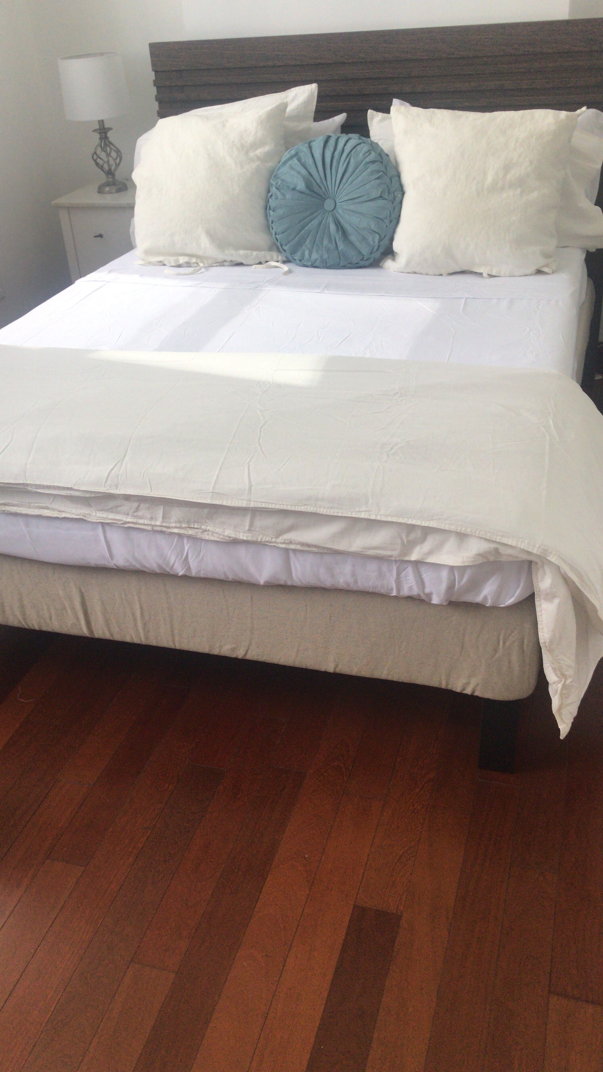 A bed with a white mattress and beige bedding, a wooden headboard, and a nightstand with a lamp.