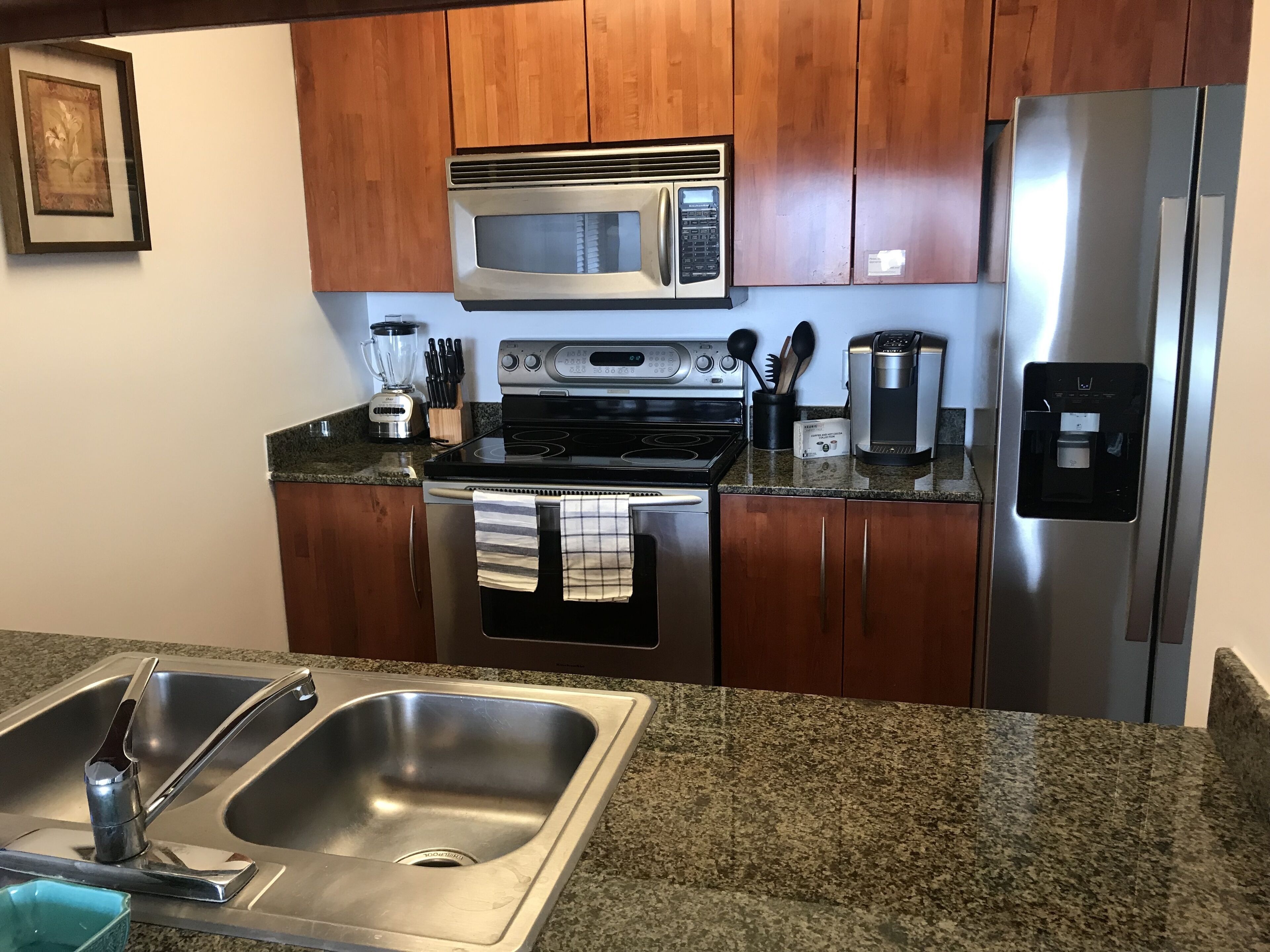 Una cocina con armarios de madera, electrodomésticos de acero inoxidable, incluyendo un microondas, horno y refrigerador, una encimera de granito y un lavamanos doble.