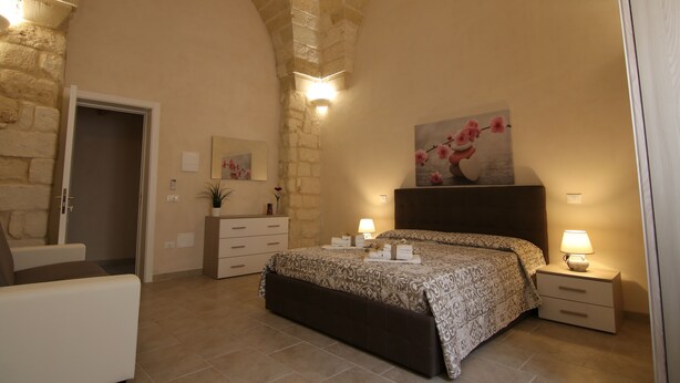 A bedroom with a stone arch ceiling, a bed with a patterned bedspread, bedside lamps, a nightstand, a dresser, and a painting on the wall. - Corte Manzoni B&B (Melendugno)