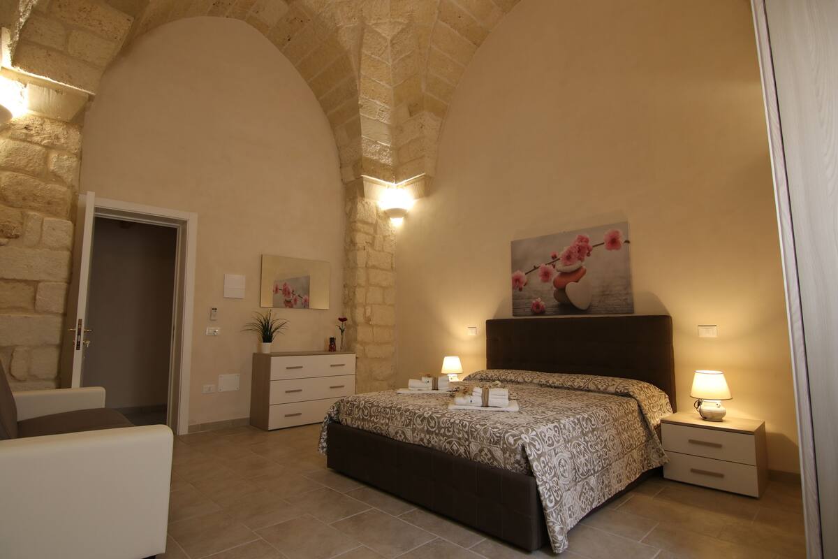 A bedroom with a stone arch ceiling, a bed with a patterned bedspread, bedside lamps, a nightstand, a dresser, and a painting on the wall.