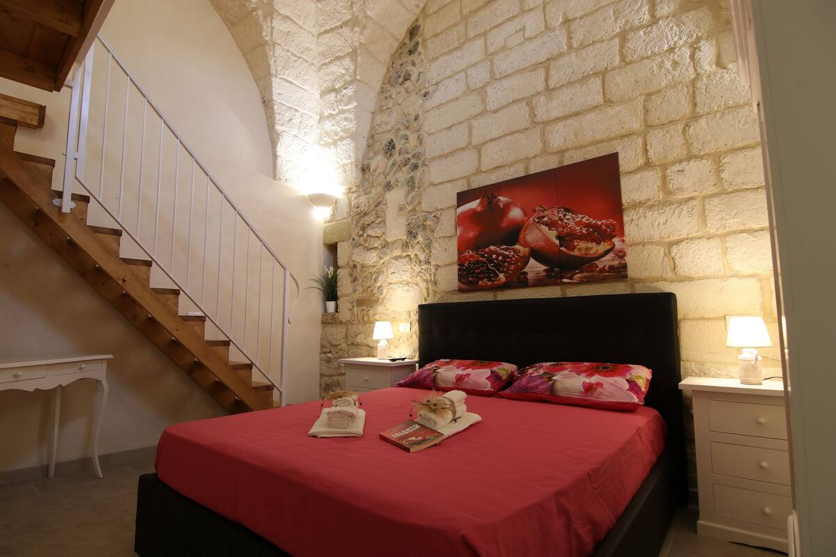 A bedroom with a red bedspread, a wooden staircase, a brick wall, and a nightstand with a lamp.
