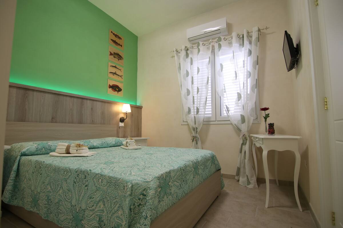 A bedroom with a bed, a bedside table, a wall-mounted air conditioner, and a window with curtains.