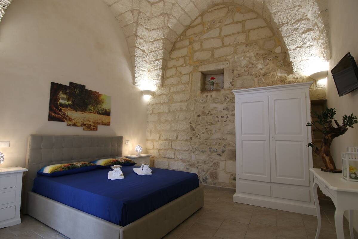 A bedroom with a stone wall, a bed with blue bedding, a white wardrobe, and a small plant.
