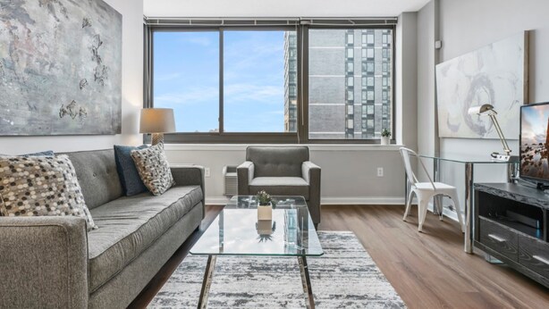 A modern living room with a grey sofa, a glass coffee table, and a view of tall buildings through large windows. - Bluebird Suites near PATH (Jersey City)