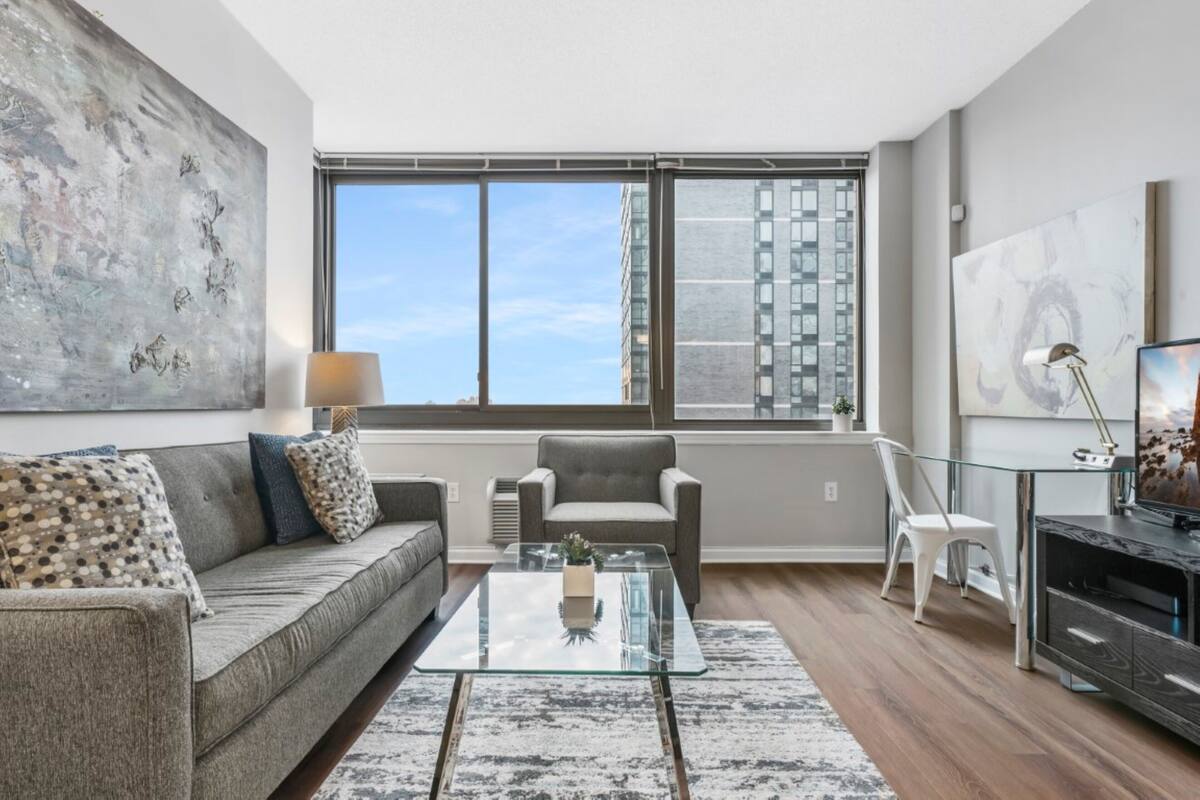 Un salón moderno con un sofá gris, una mesa de centro de cristal y vistas a edificios altos a través de grandes ventanales.
