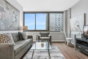 Un salón moderno con un sofá gris, una mesa de centro de cristal y vistas a edificios altos a través de grandes ventanales.
