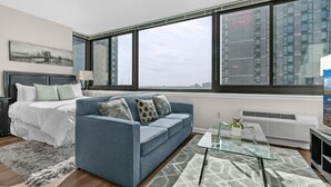 A modern bedroom with a bed, a blue sofa, a glass coffee table, and large windows offering a city view. - Bluebird Suites near PATH (Jersey City)
