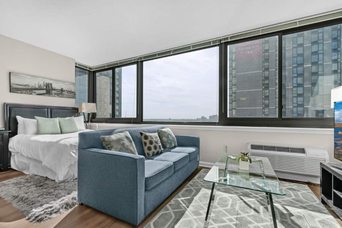 Un dormitorio moderno con una cama, un sofá azul, una mesa de centro de vidrio y ventanas amplias con vistas a la ciudad.