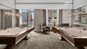 A modern hotel room with two pool tables, a comfortable armchair, and a large window offering a city view. - Bluebird Suites near PATH (Jersey City)