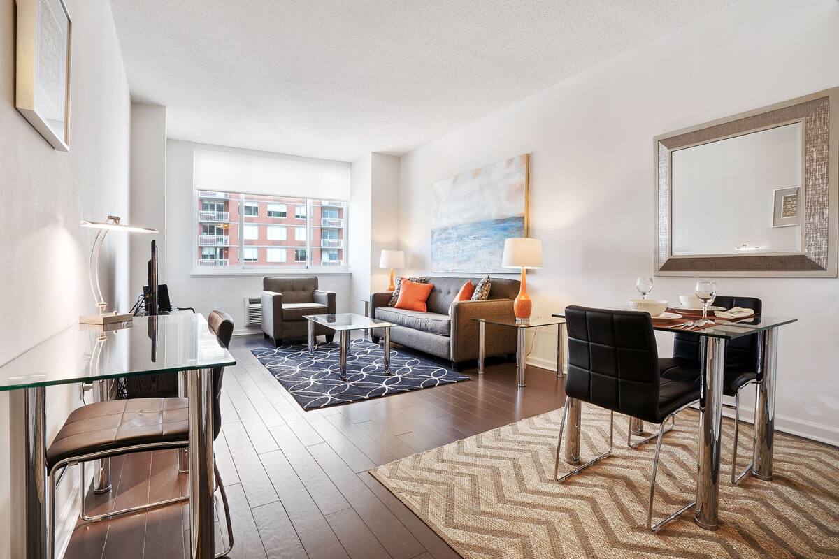 Ein modernes Wohnzimmer mit einem Glastisch, Ledersesseln, einer Couch mit orangefarbenen Kissen und einem großen abstrakten Gemälde an der Wand.