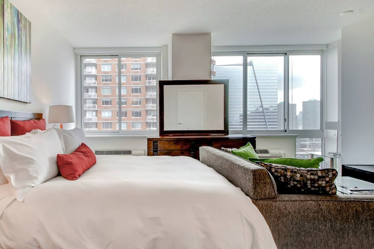 Ein Hotelzimmer mit einem großen Bett, einem Fernseher, einer Couch und Blick auf eine Stadtlandschaft durch große Fenster.