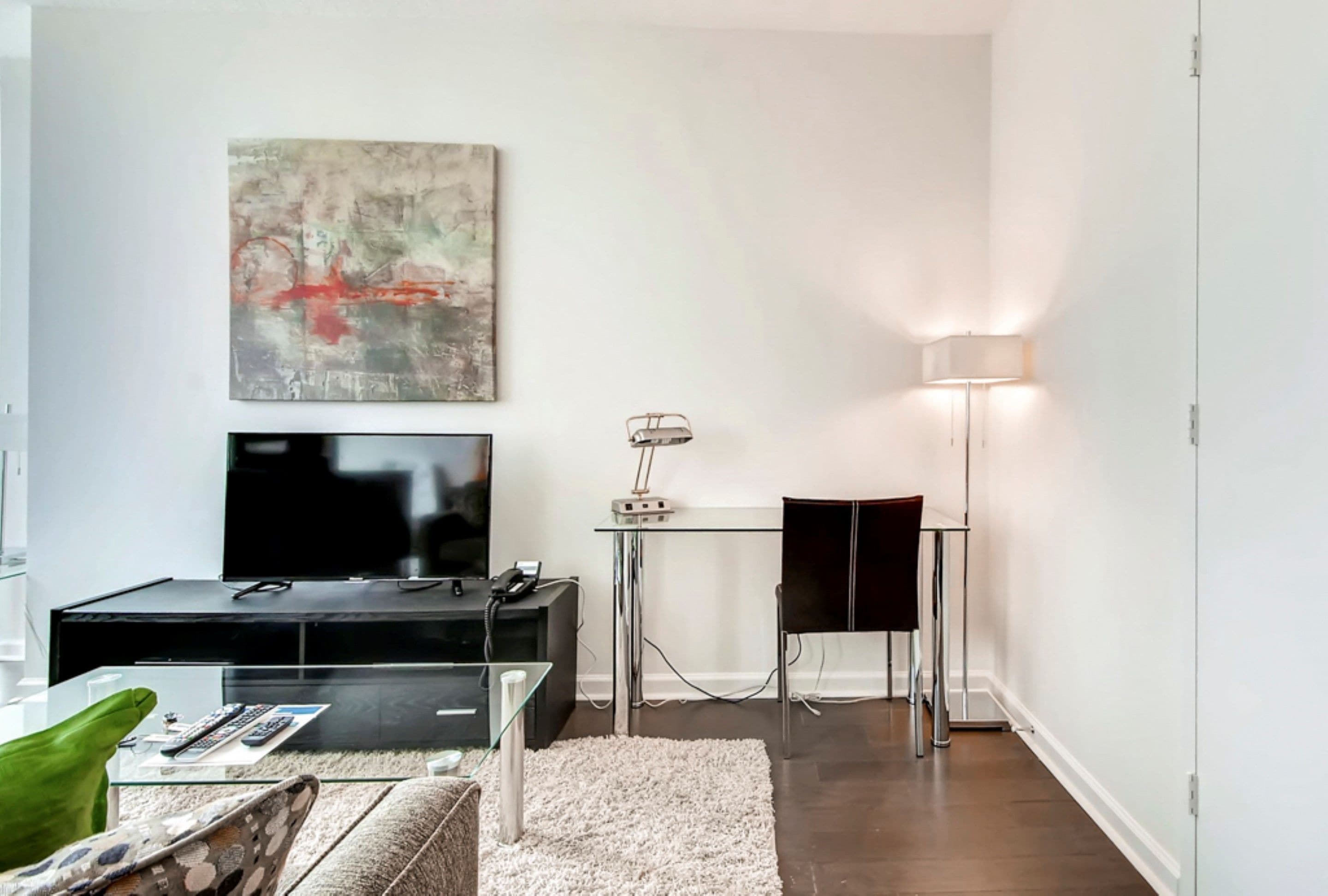 A modern living room with a flat-screen TV, a glass coffee table, a sofa, a small desk with a lamp, and a painting on the wall.