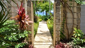 A stone pathway leading to a wooden archway, surrounded by lush greenery and a view of the ocean. - Secret Cove 1 by Altman (Paynes Bay)