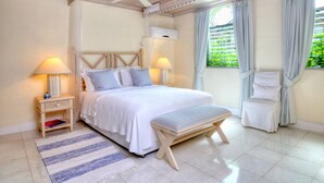 A bedroom with a bed, bedside tables, a bench, a ceiling fan, and a window with curtains. - Secret Cove 1 by Altman (Paynes Bay)